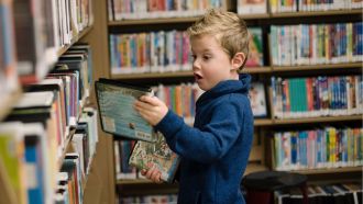 Young boy pulls books off shelf in library.