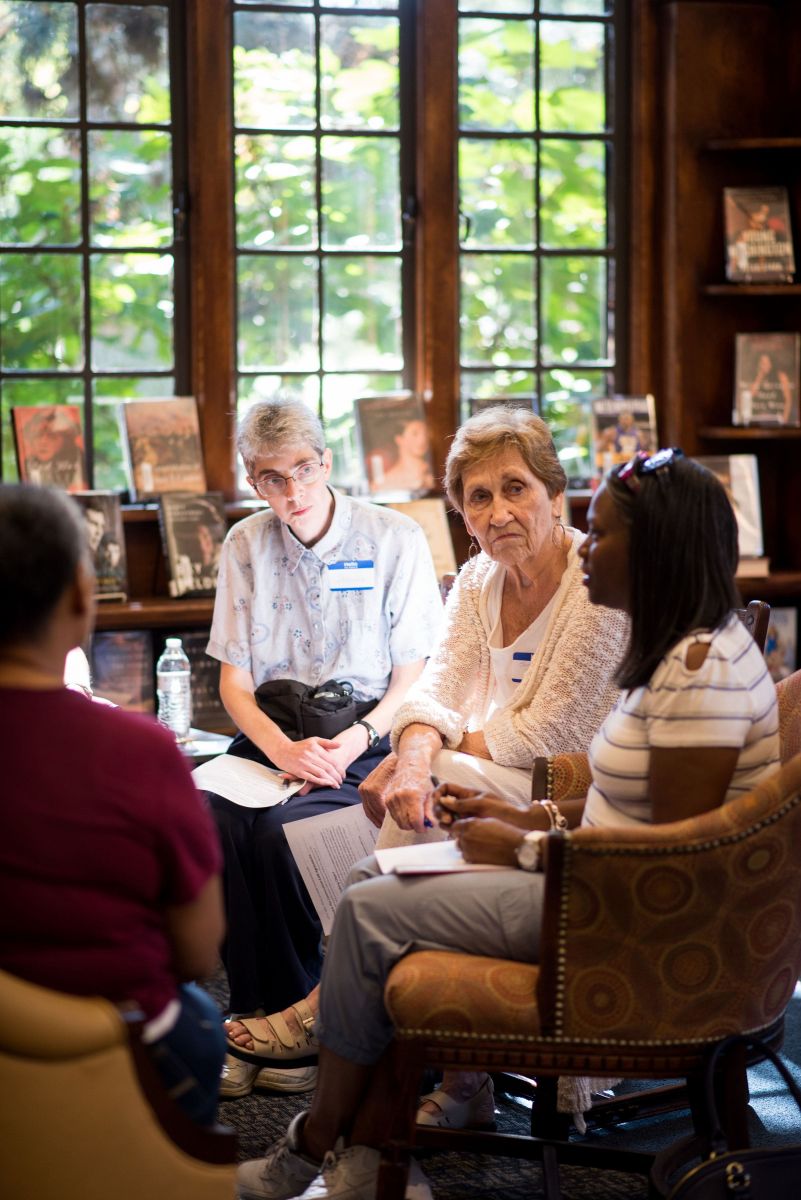 a young black woman speaks as older white women listen in front of a sunlit library window